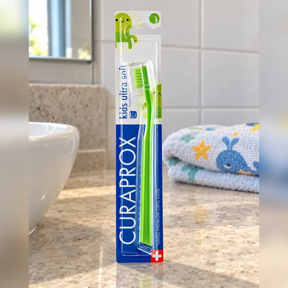 Escova Curaprox Kids verde na cartela azul ultra soft, em pé na bancada clara do banheiro, com uma toalha infantil de baleia dobrada ao fundo
